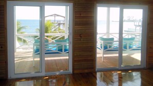 Ein Zimmer mit Holzwänden und Holzboden, große Schiebeglasfenster zu einem Außendeck mit blauen Stühlen und Blick aufs Meer.