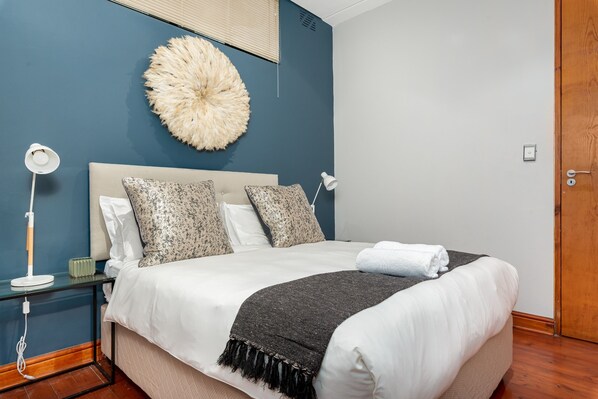 Un lit bien fait, avec une couette blanche et un plaid foncé, une décoration murale moelleuse, une lampe de chevet et une table de chevet sur laquelle se trouve un objet vert.
