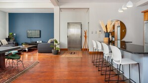 Ein modernes Wohnzimmer mit Sofa, Couchtisch und Sesseln. Es gibt eine Essecke mit einer Bar und Stühlen. Ein Fernseher ist an der Wand montiert und eine Pflanze steht auf dem Boden.