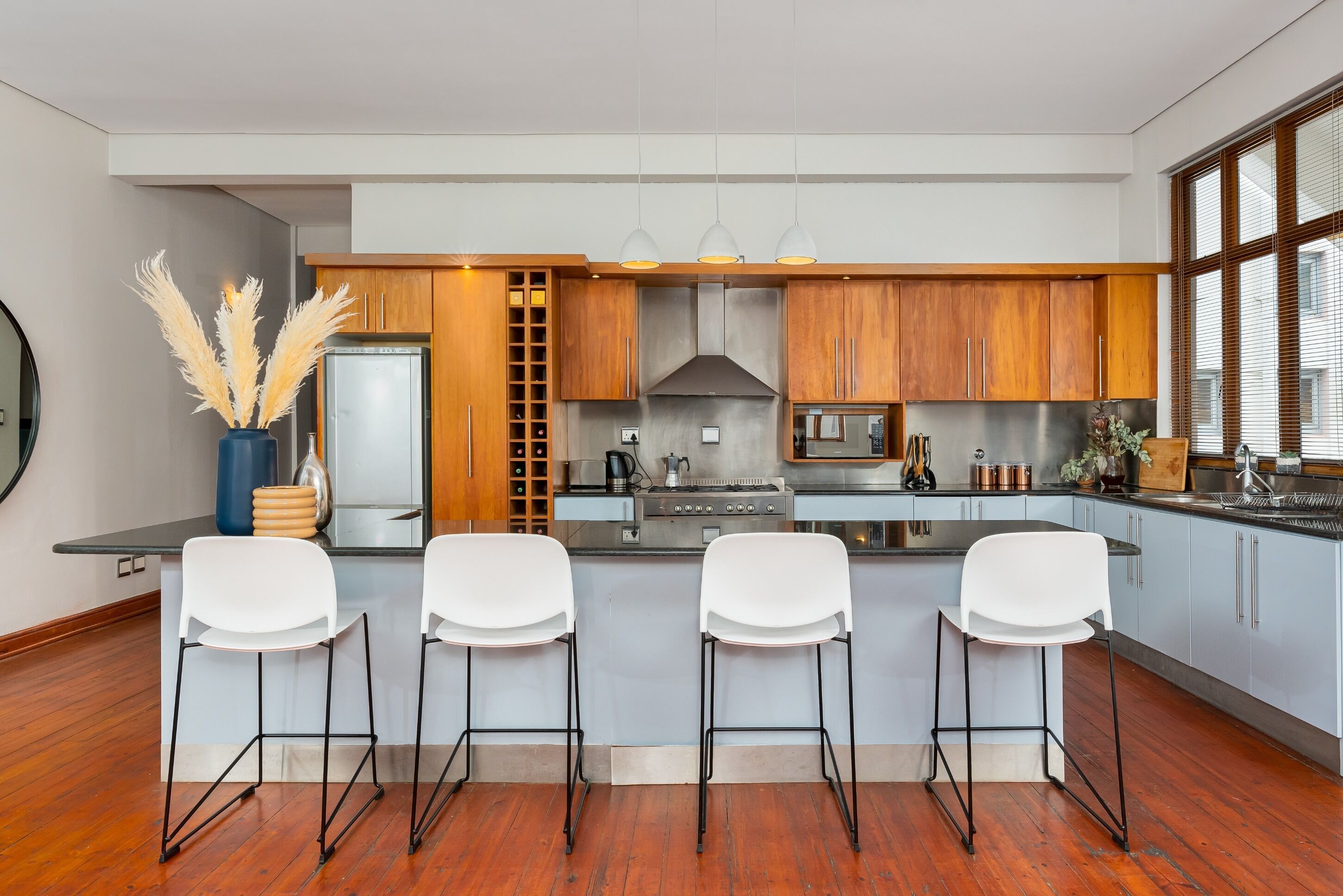 A modern kitchen with wooden cabinets, a central island with bar stools, stainless steel appliances, and a large window providing natural light.