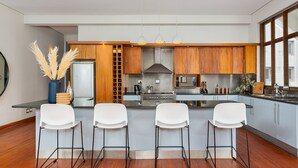 Eine moderne Küche mit Holzausstattung, einer zentralen Kochinsel mit Barhocker, Edelstahlgeräten und einem großen Fenster, das für natürliches Licht sorgt.