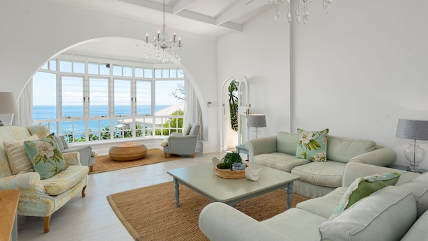 Amplio salón con vistas al mar, que cuenta con un gran ventanal en arco, elegantes lámparas de araña y muebles de colores claros.