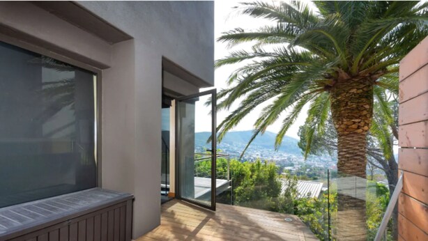 A balcony with a wooden floor, a glass door leading to a city view, and a palm tree. - Nature Lover's Nest at the Foot of Table Mountain (Cape Town)