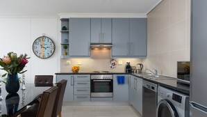 Une cuisine moderne avec des meubles gris, un four en acier inoxydable, un lave-linge et un coin repas agrémenté d’un vase de fleurs.