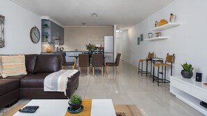 Un salon moderne avec un canapé en cuir marron, un coin repas doté d’une table en verre et une cuisine en arrière-plan.