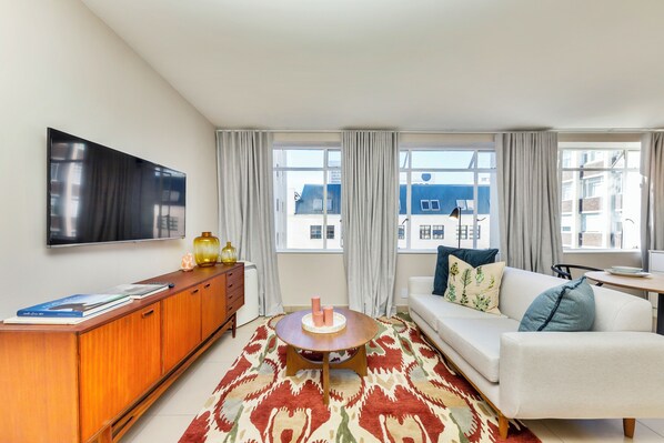 A modern living room with a sofa, a coffee table, a TV mounted on the wall, and large windows with curtains. - Refurbished Studio Apart in the Heart of Cape Town
(Cape Town)