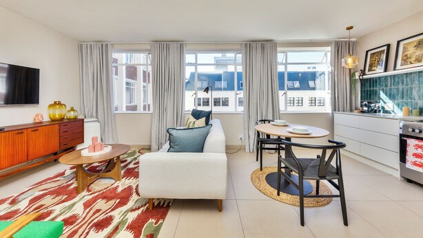 A modern living room with a sofa, coffee table, and dining area. There is a kitchen with a stove and a TV mounted on the wall. - Refurbished Studio Apart in the Heart of Cape Town

 (Cape Town)
