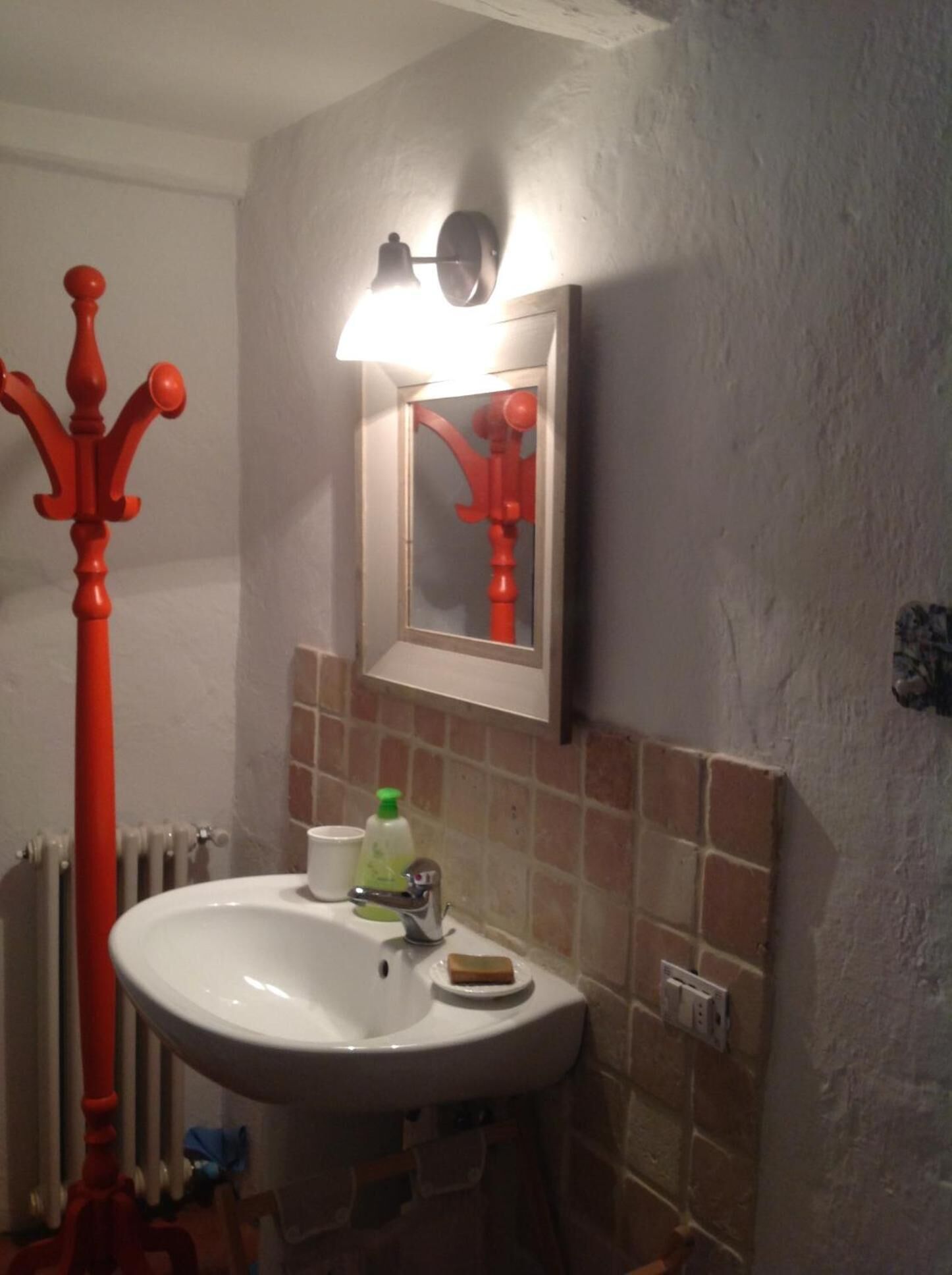 A bathroom with a white sink, a red coat rack, a mirror with a red reflection, and a wall-mounted light fixture.