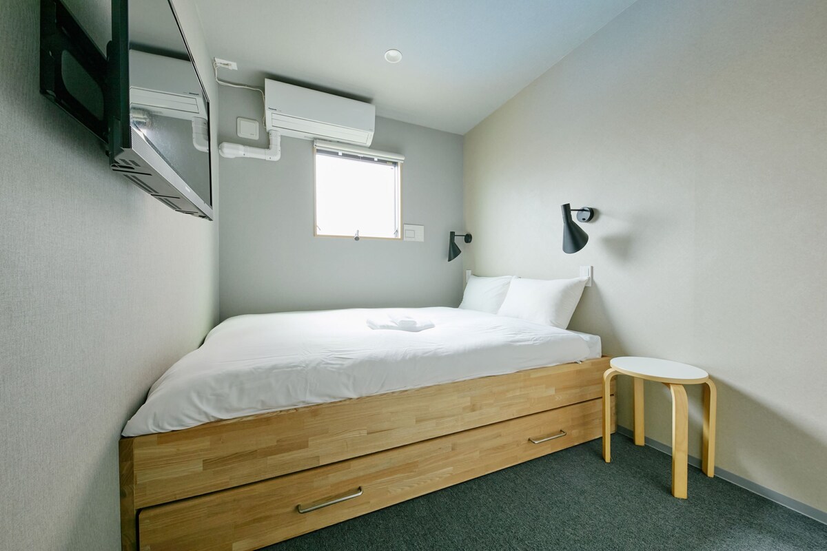 A bedroom with a wooden bed, a small stool, a wall-mounted lamp, and an air conditioning unit.