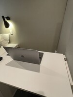 Un ordenador portátil sobre un escritorio blanco, con una lámpara de pared y un cojín blanco.