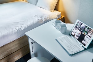 Una cama bien hecha con ropa blanca, un escritorio blanco con una computadora portátil y dos tazas blancas, y una mesita de noche con una lámpara.