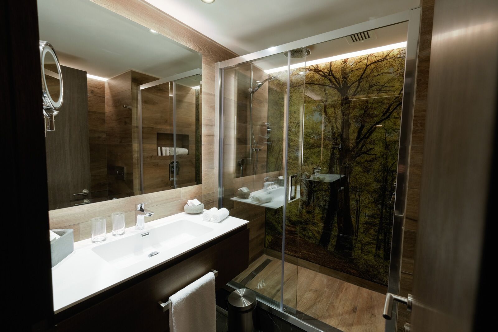 A modern bathroom with a large mirror, a white sink, and a glass shower enclosure with a forest mural.