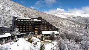 A large, multi-story building with a snow-covered roof, surrounded by snow-laden trees and mountains. - Hotel Termas Chillán (Pinto)