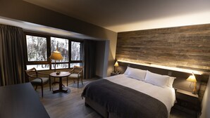 A modern hotel room with a large bed, a desk, two chairs, a small table, and a view of trees through the window. - Hotel Termas Chillán (Pinto)