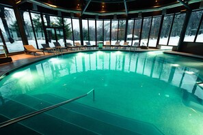 A large indoor swimming pool with clear blue water, surrounded by lounge chairs and large windows offering views of a snowy landscape.