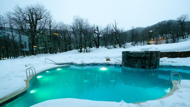 A swimming pool surrounded by snow with a stone wall and metal ladders. - Hotel Termas Chillán (Pinto)