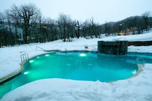 A swimming pool surrounded by snow with a stone wall and metal ladders. - Hotel Termas Chillán (Pinto)