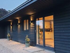 A modern house with a dark exterior, large sliding glass doors leading to a kitchen area, and two potted plants on the gravel driveway. - Chicken Coop, Cottage, Fishing Lakes Nr Cleethorpes, Adults Only Pet Friendly (Lincolnshire)
