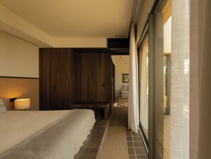 A hotel room with a bed, a nightstand, a lamp, a wooden wardrobe, and a sliding glass door leading to a balcony.