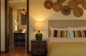Habitación de hotel con cama, mesita de noche con lámpara, baño con lavamanos y espejo, y decoración en la pared.