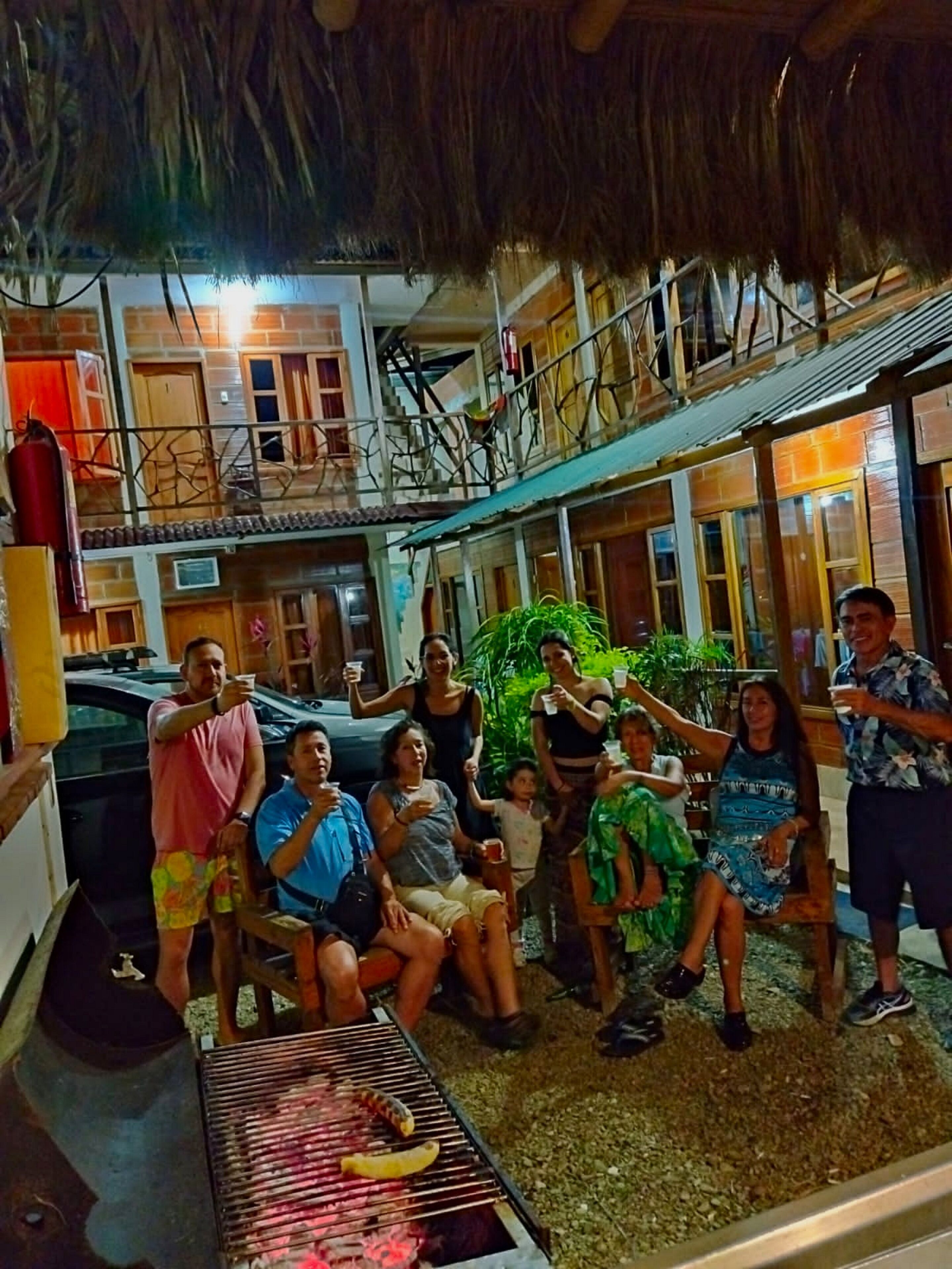 Un grupo de personas reunidas alrededor de una parrilla con comida, sentadas en sillas y bancos, en un espacio al aire libre con techo de paja y construcciones de madera.