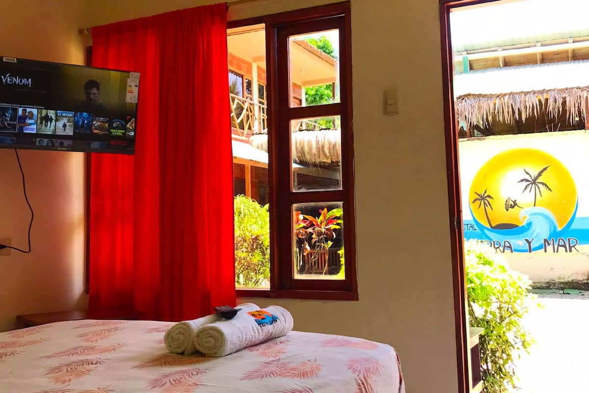 A bedroom with a bed, a TV mounted on the wall, a window with red curtains, and a view of a beach.