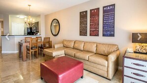 Un salon avec un canapé d’angle beige, un pouf rouge, un coin repas avec des chaises et une table en bois, et une cuisine en arrière-plan.