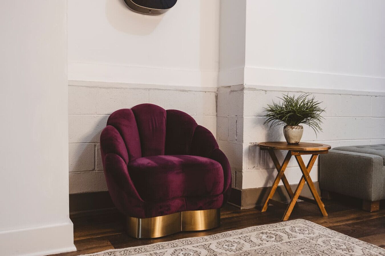 A corner room with a dark red velvet armchair, a small wooden side table with a plant, and a grey upholstered bench.