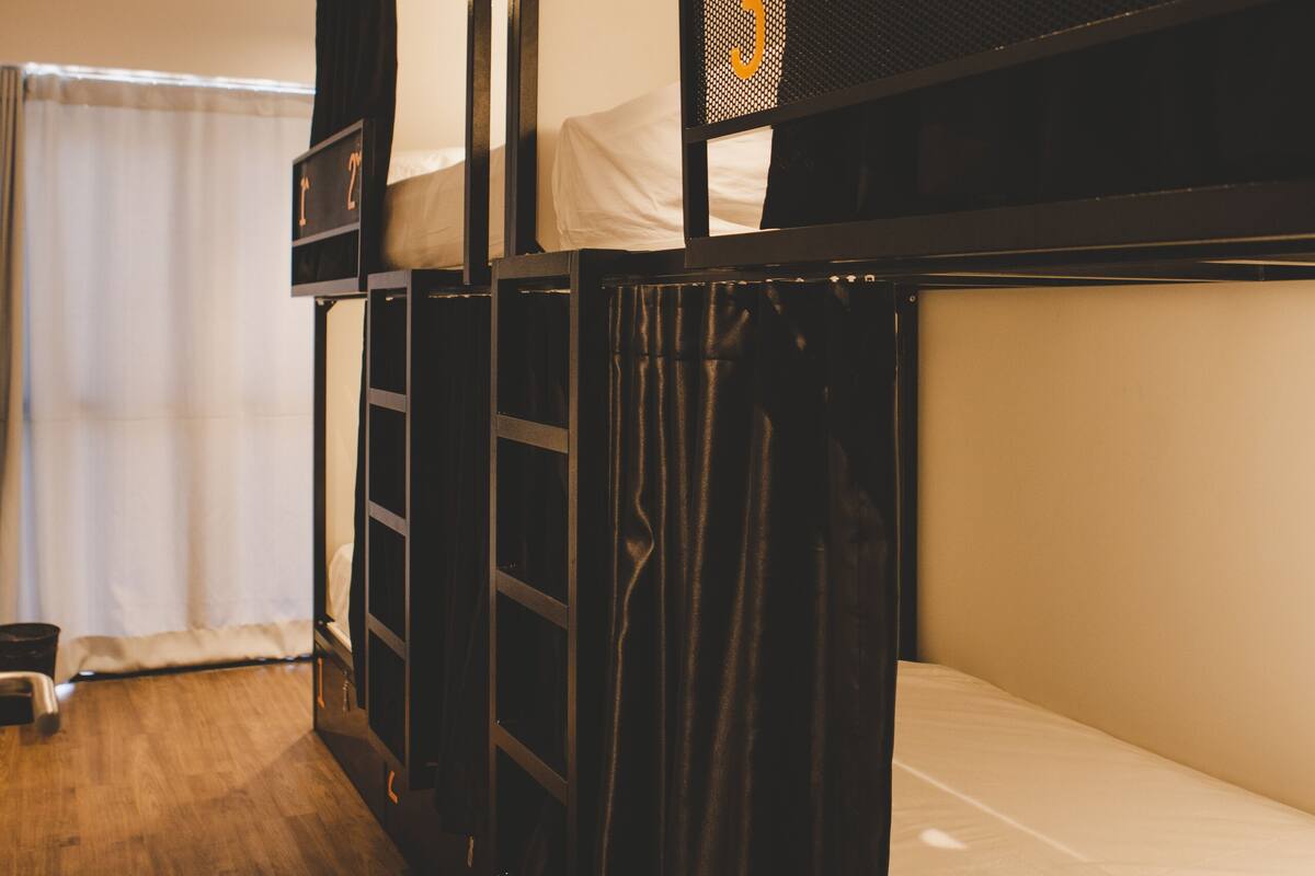 Quarto com beliche, uma mesinha e janela com cortinas.