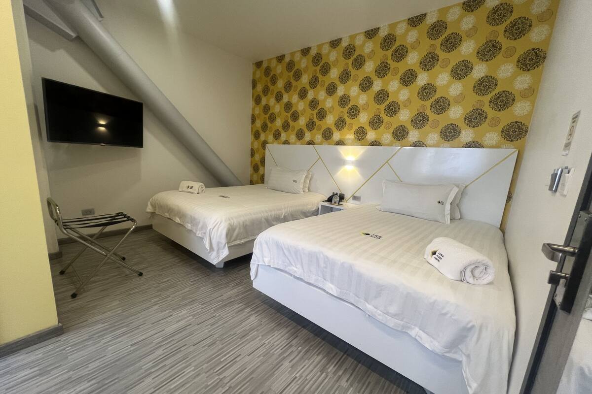 A hotel room with two beds, a flat-screen TV mounted on the wall, a small table with a lamp, and a chair.
