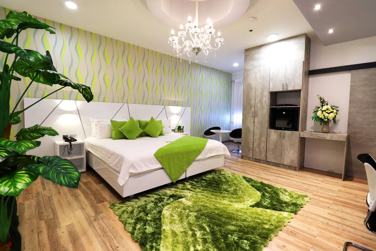 A modern bedroom with a bed, green pillows, a chandelier, a desk, and a plant.