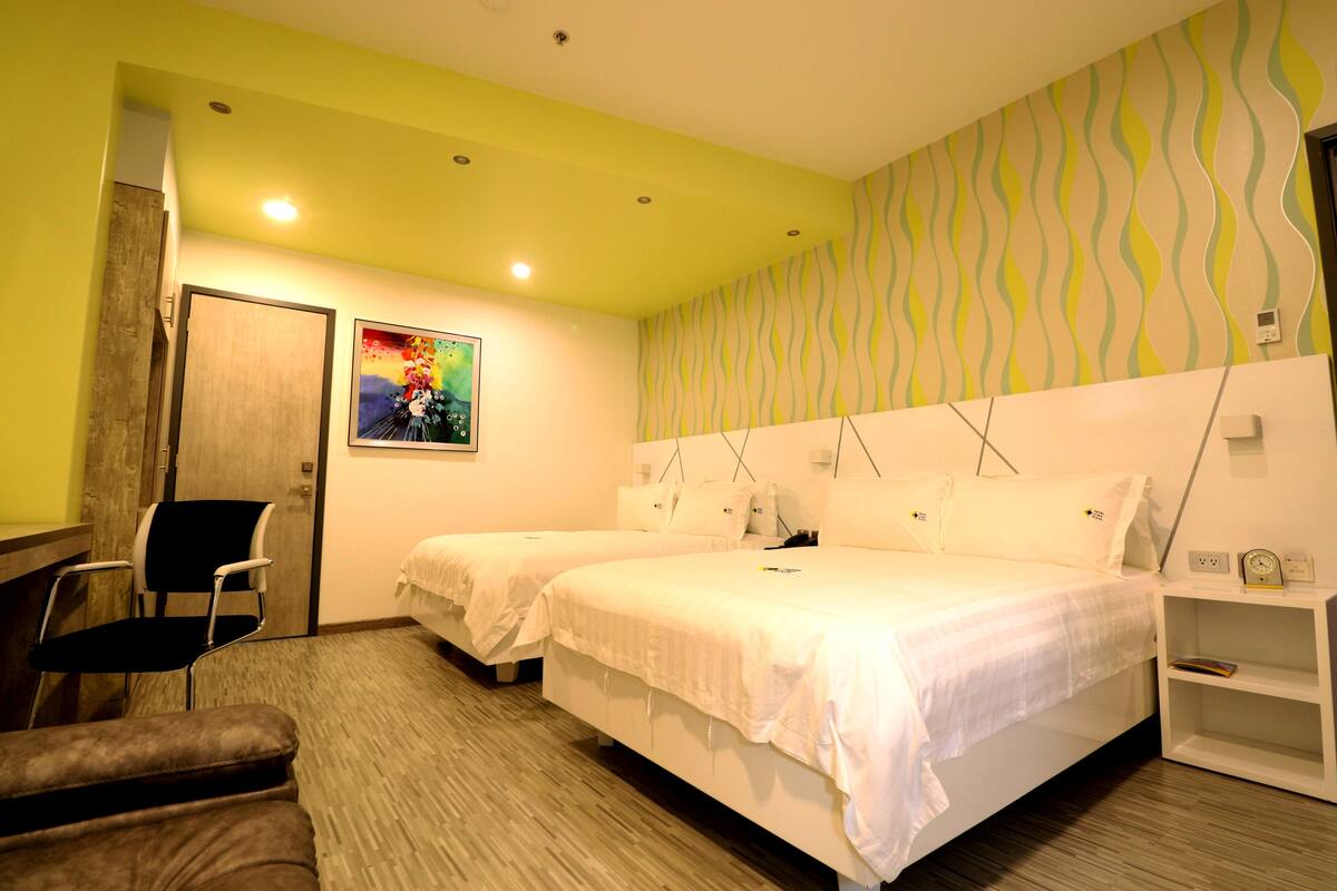 A hotel room with two single beds, a desk, a chair, and a small shelf with a clock.