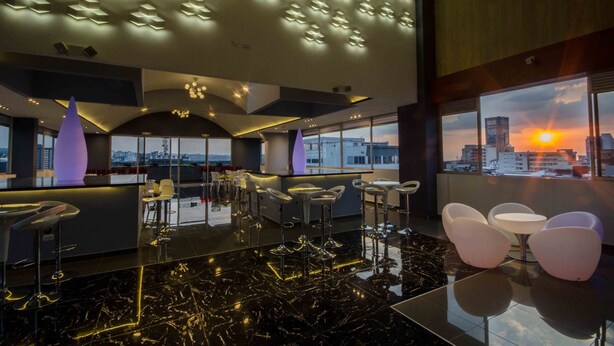 Un área de bar moderna con vistas a la ciudad al atardecer, que presenta una barra elegante, taburetes altos y un suelo de mármol pulido.