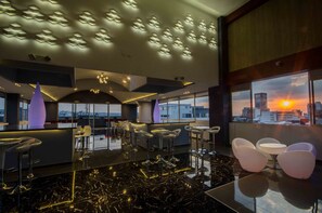 Un área de bar moderna con vistas a la ciudad al atardecer, que presenta una barra elegante, taburetes altos y un suelo de mármol pulido.