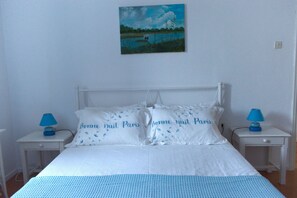 A neatly made bed with white linens and two pillows with text, a small bedside table with a lamp, and a painting on the wall.