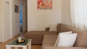 Ett vardagsrum med en beige soffa, ett litet bord med en flaska och ett ljus, och en tavla med blommor på väggen.