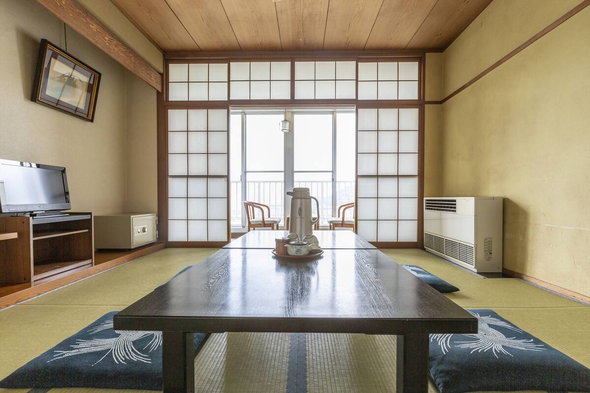 A traditional Japanese room with tatami flooring, a low table with a tea set, a television, and a sliding door with a view to the outside.