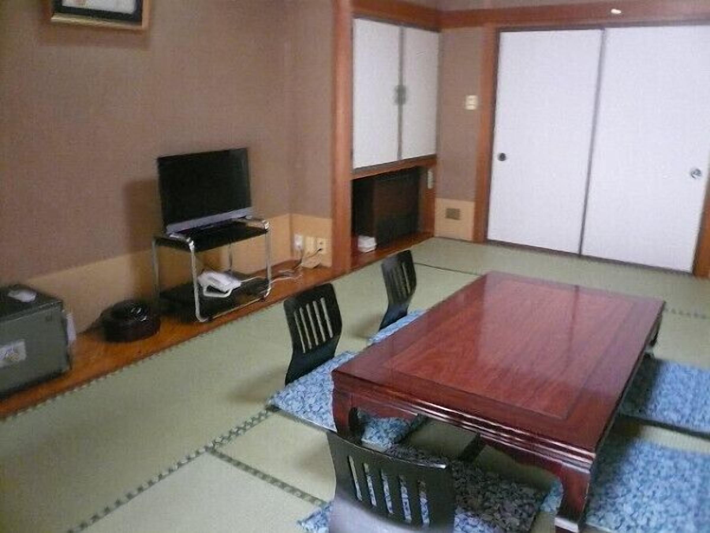 A room with a wooden table, chairs, a television on a stand, a small shelf, and a wardrobe.