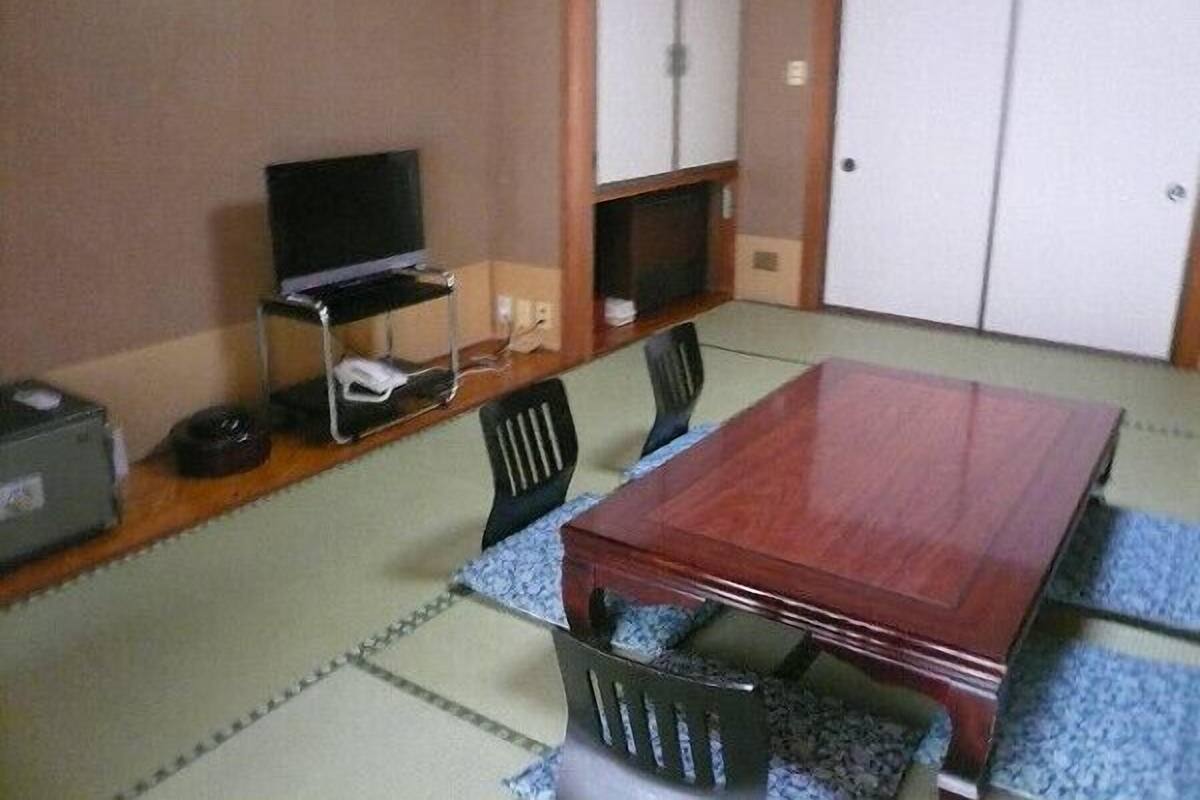 A room with a wooden table, chairs, a television on a stand, a small shelf, and a wardrobe.