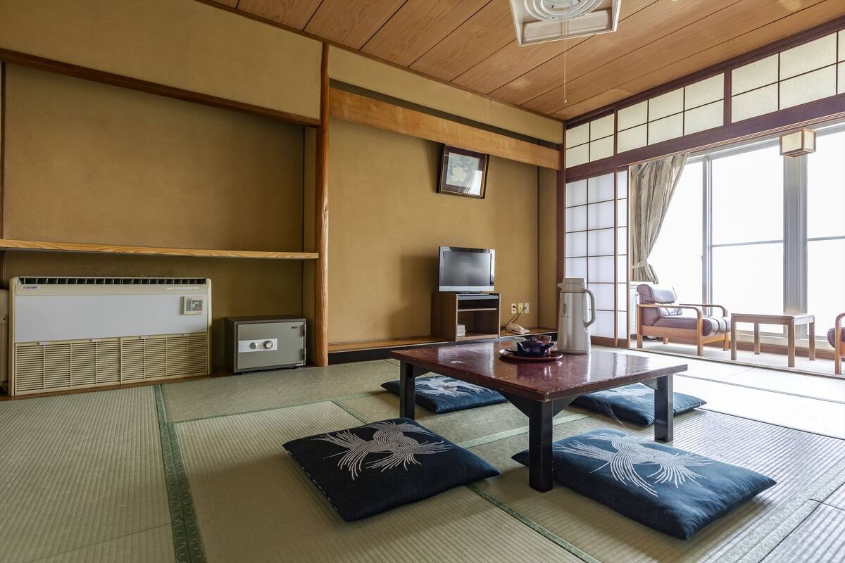 A traditional Japanese room with tatami flooring, a low table with cushions, a television, and a sliding door to a balcony.