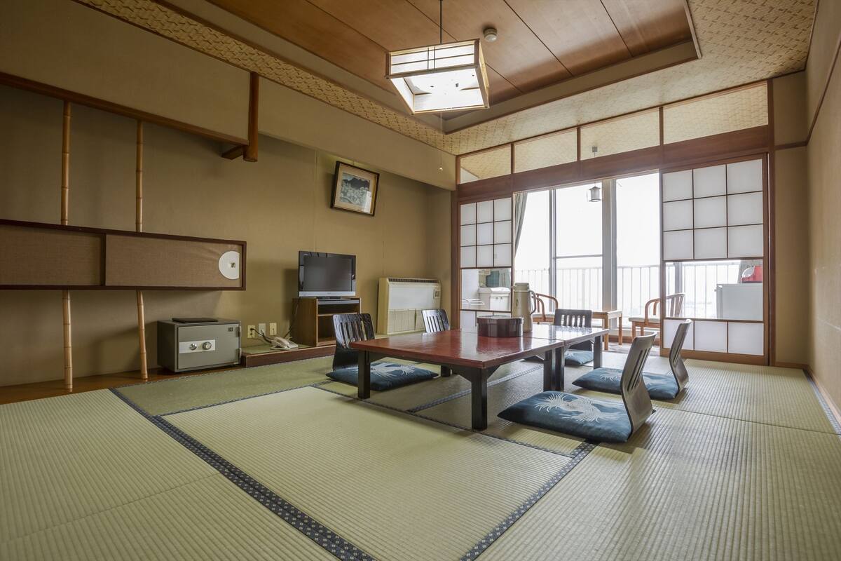 A traditional Japanese room with tatami flooring, a low table with cushions, a television, and a sliding door leading to a balcony.