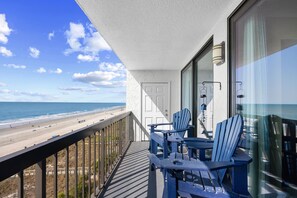 A balcony with a view of the beach, a door, and two Adirondack chairs.