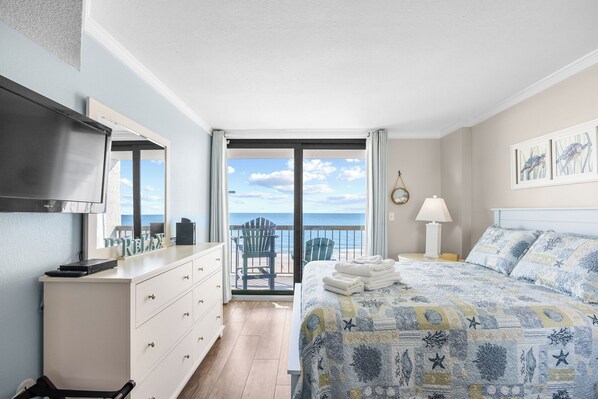 A bedroom with a bed, a dresser, a TV, a balcony with a chair, and a view of the ocean.