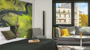 A modern hotel room with a large window, a bed with a green cover, a grey sofa, and a small round table with a book and a mug. - Novotel Paris Porte De Versailles (Paris)