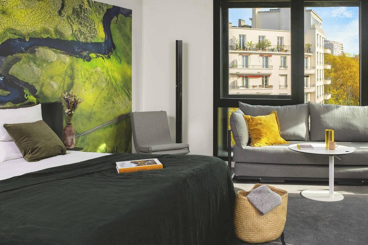 A modern hotel room with a large window, a bed with a green cover, a grey sofa, and a small round table with a book and a mug.