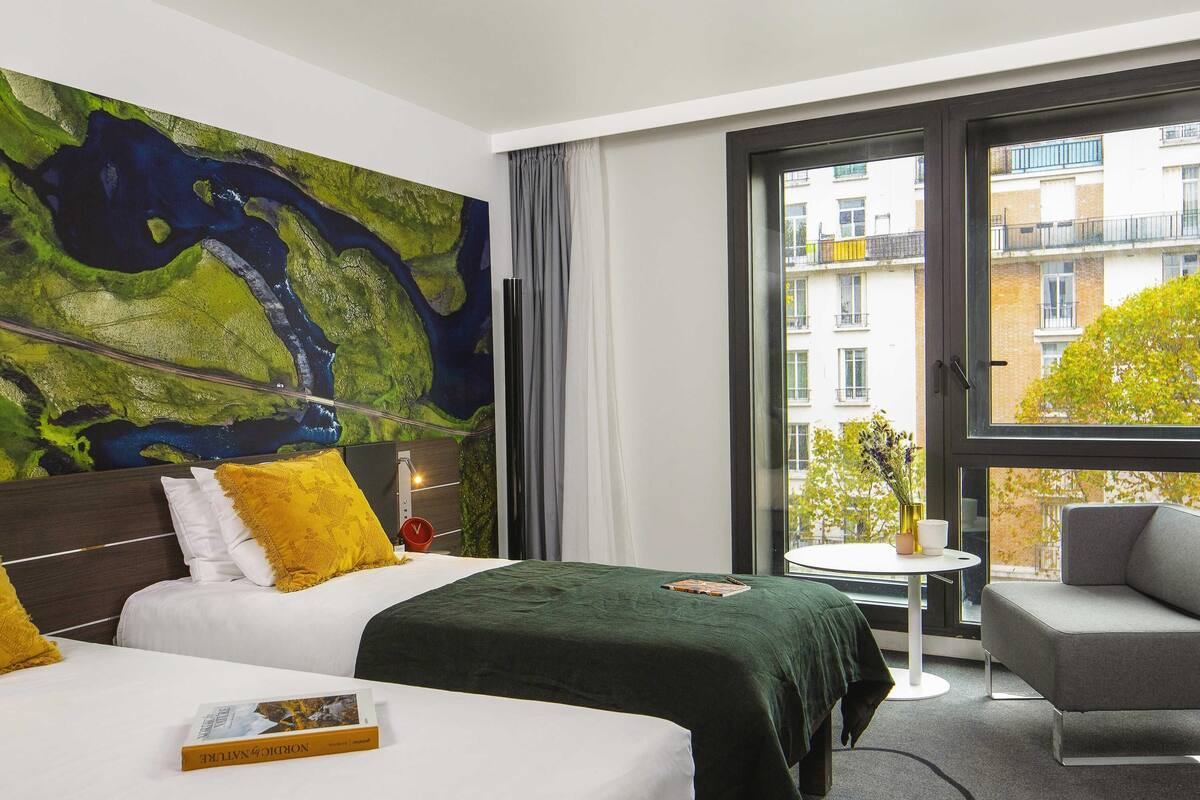 A hotel room with a large window, a bed with a green bedspread, a grey armchair, and a small round table.