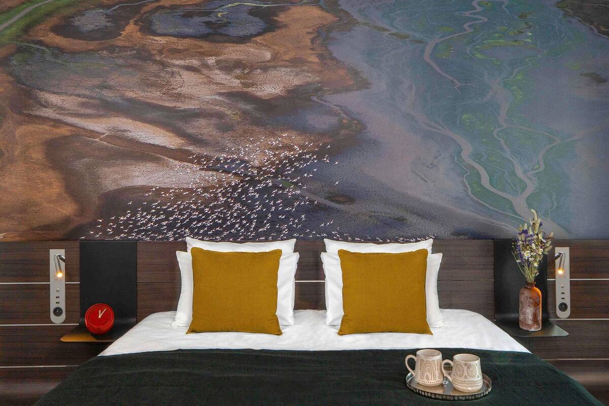A bed with two pillows, a clock, a vase with flowers, and a tray with two mugs.