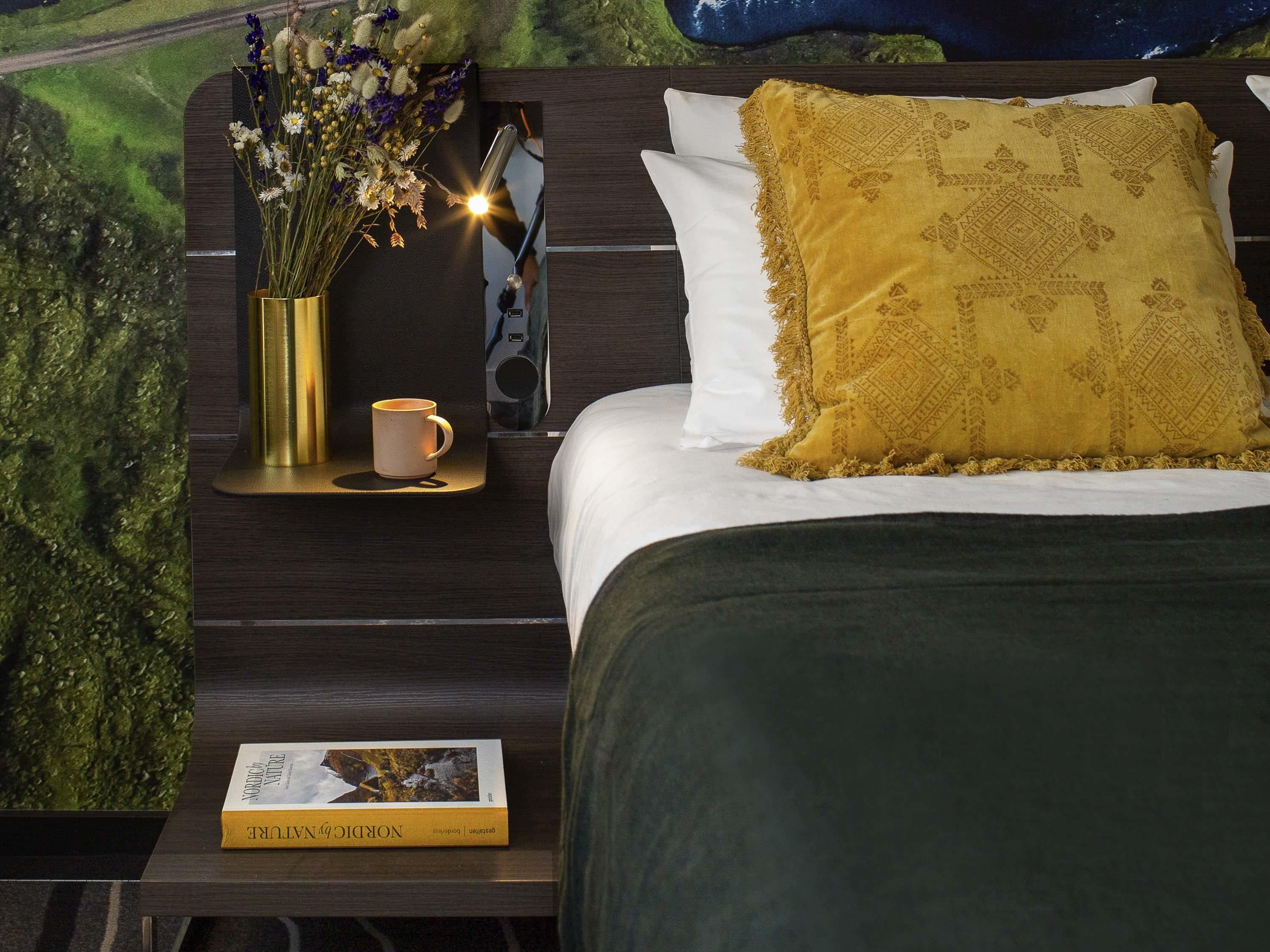 Cama bem arrumada com almofada estampada, mesa de cabeceira com um livro e uma xícara, e luminária fixada na parede.