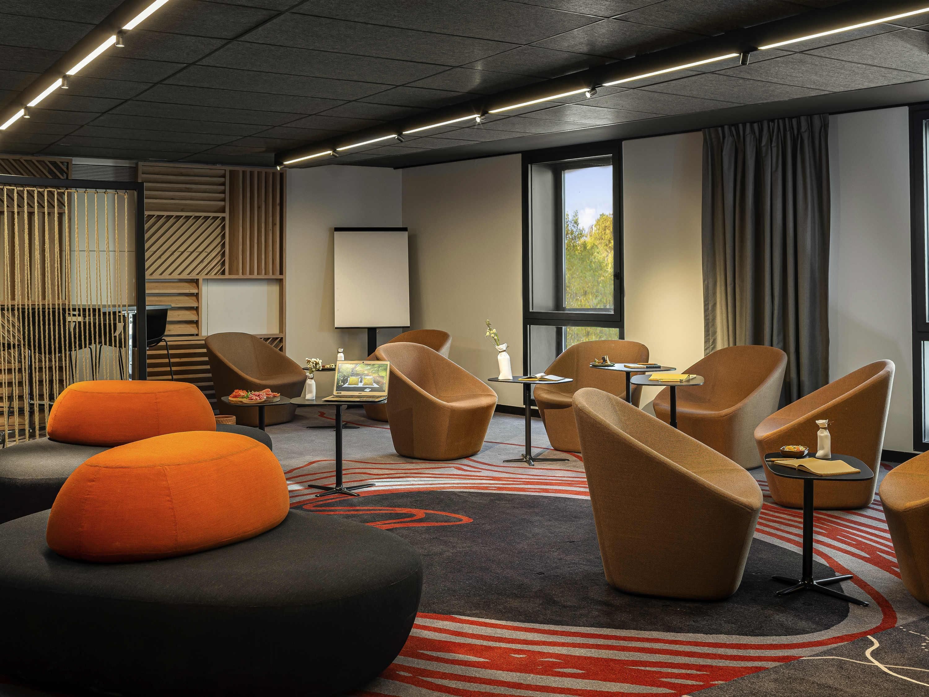 A hotel room with a modern design, featuring a large window, a flat-screen TV, and multiple seating areas with orange and black chairs.