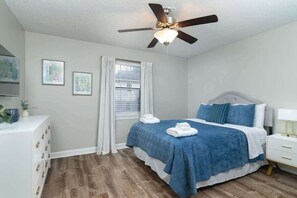 Quarto com cama, ventoinha no teto, janela com cortinas, mesa de cabeceira e cômoda.